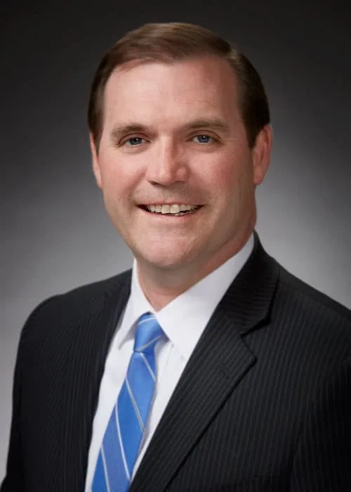 Man in dark pinstriped suit and blue tie smiling at camera against gray background