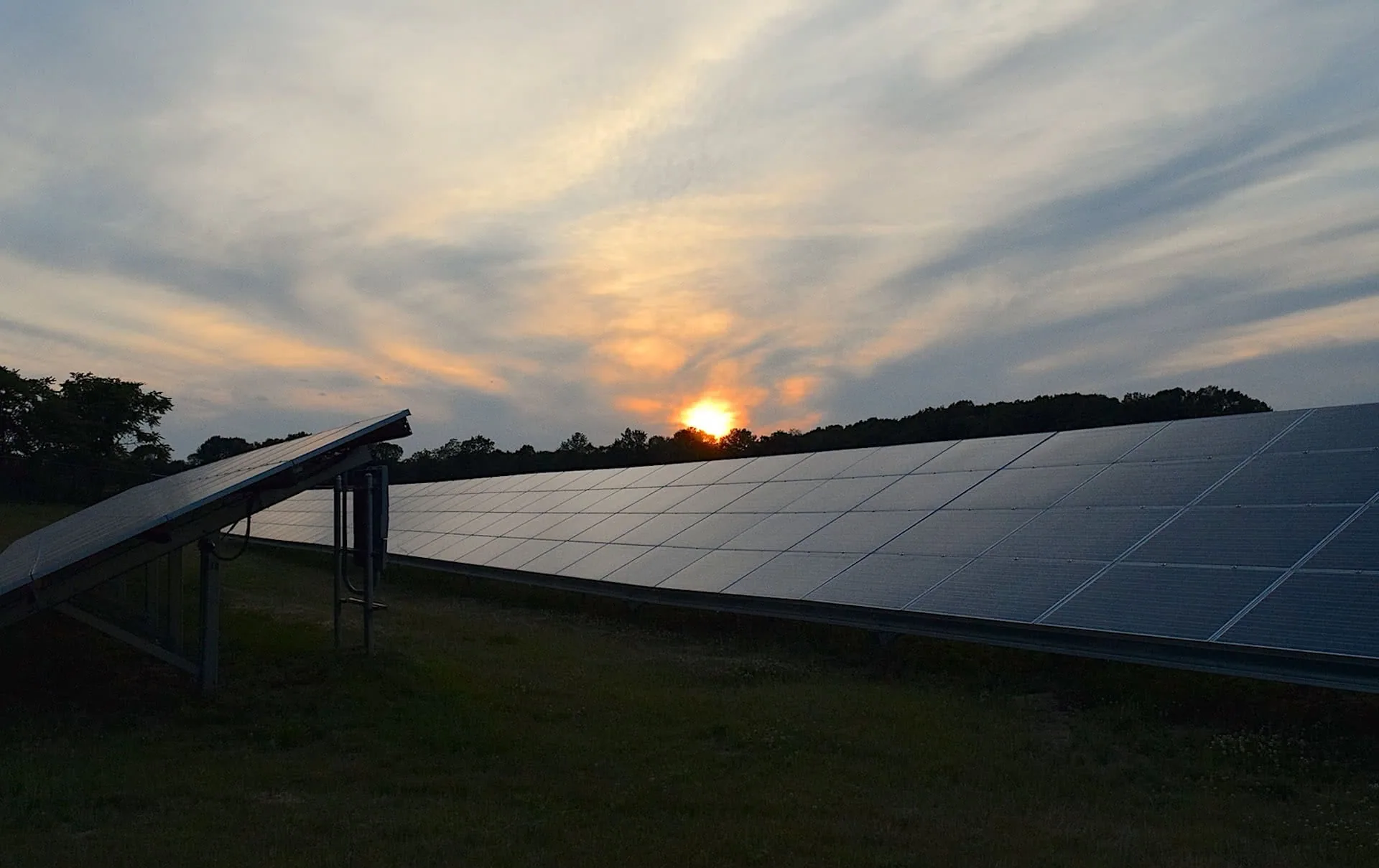 Solar panel array on grass field at sunset with trees silhouetted against orange and blue sky